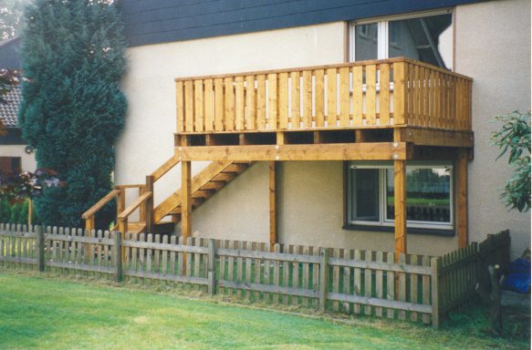 Holzterrasse mit Treppe neben einem Haus, umgeben von einem niedrigen Zaun.